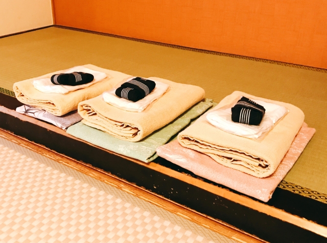 Neatly arranged folded blankets and futons on tatami flooring, with individual black and white slippers on top.