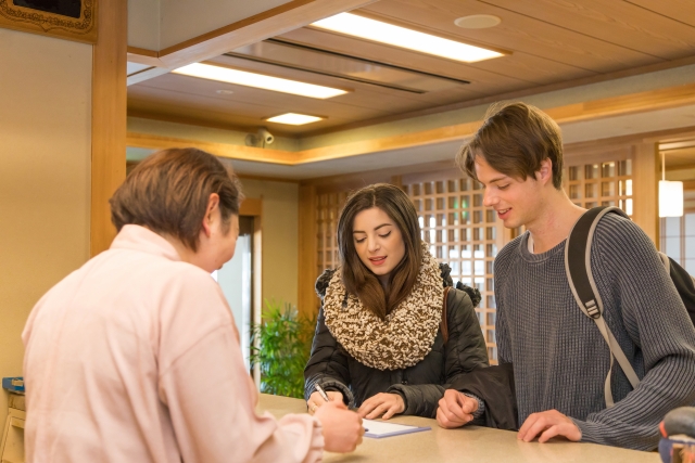A woman at a reception desk assisting two young travelers, one of whom is signing in, in a warm and welcoming environment.