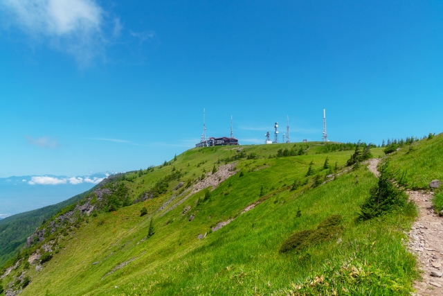 Panoramic view of a green hillside with communication towers at the summit under a clear blue sky.